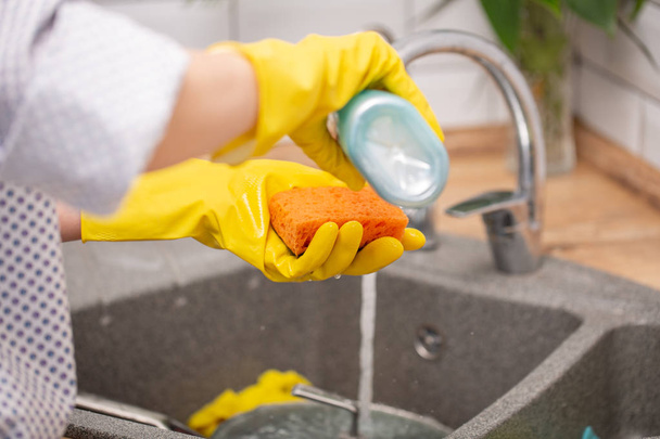 Домохозяйка в резиновых перчатках моет посуду с оранжевой губкой на кухне дома
. - Фото, изображение