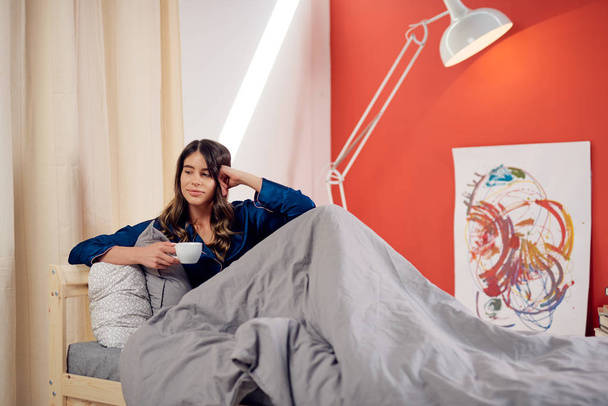 Pretty Caucasian brunette sitting on bed in bedroom, smiling and drinking morning coffee. - Photo, Image