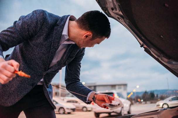 Jonge manager formeel gekleed controleren de olie van zijn auto op zonsondergang wh - Foto, afbeelding