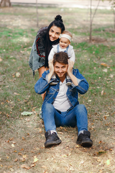 A happy father is sitting on a lawn, a daughter is on his shoulders, a mother is hugging a family, a beautiful autumn day, a beautiful stylish family - Photo, image