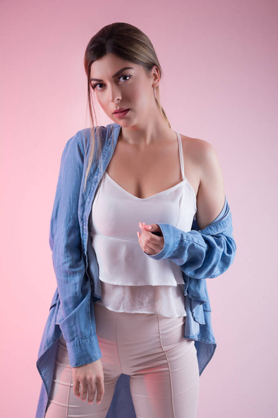 Uwodzicielski, młoda kobieta z barkiem gołej nosi niebieskie dżinsy koszula na białym tle na różowym tle w studio. Dziewczyna patrząc na kamery. Koncepcja moda portret. Z bliska - Zdjęcie, obraz