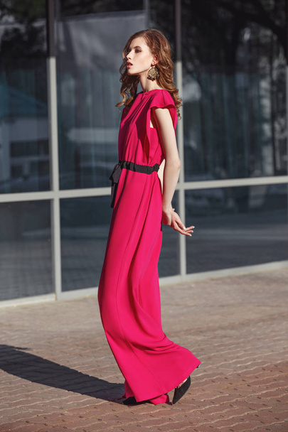 Slim stylish brunette girl dressed in fuchsia color overalls is walking in the city street on a sunny day - Fotó, kép