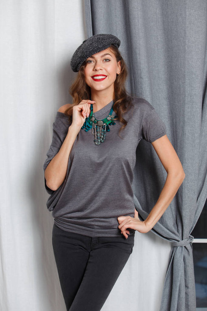 Fashionable girl dressed in a stylish gray blouse with green beads, gray jeans and a stylish cap poses in the studio - Zdjęcie, obraz