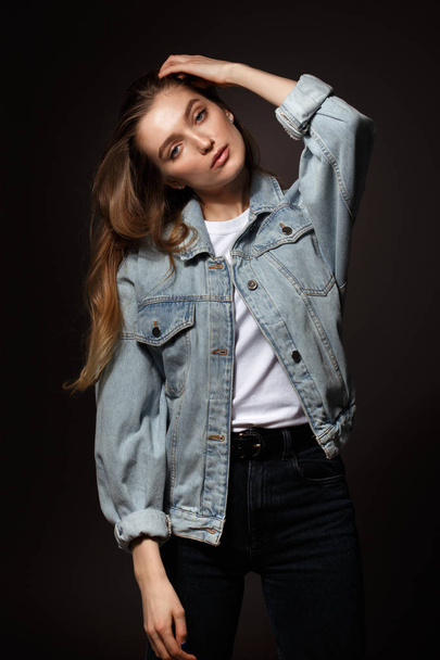 Gorgeous brunette girl with long flowing hair dressed in jeans jacket and jeans poses standing on the dark background in the studio - Fotoğraf, Görsel