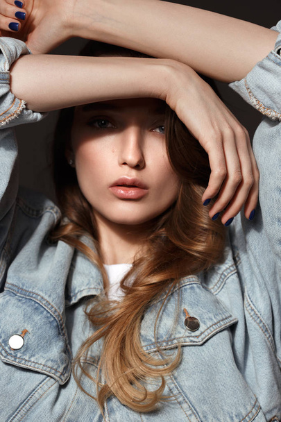 Young brunette girl with long flowing hair dressed in jeans jacket poses holding her hands on her head on the dark background in the studio - Photo, Image