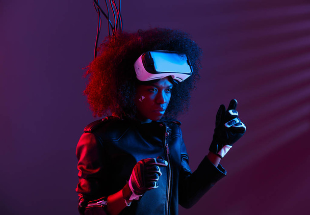 Stylish curly dark haired girl dressed in black leather jacket and gloves is wearing the virtual reality glasses on her head in the dark studio with neon light - Foto, imagen
