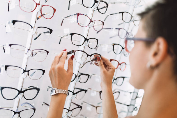 Beautiful middle age woman choosing a glasses in optician store. - Zdjęcie, obraz
