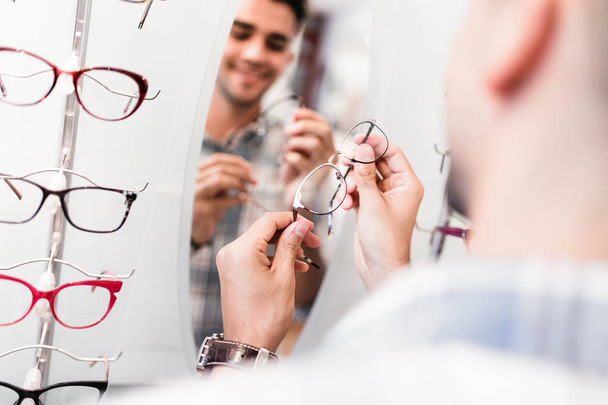 Handsome young man choosing eyeglasses frame in optical store. - Foto, Imagen