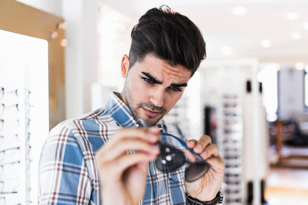 Handsome young man choosing eyeglasses frame in optical store. - Fotó, kép