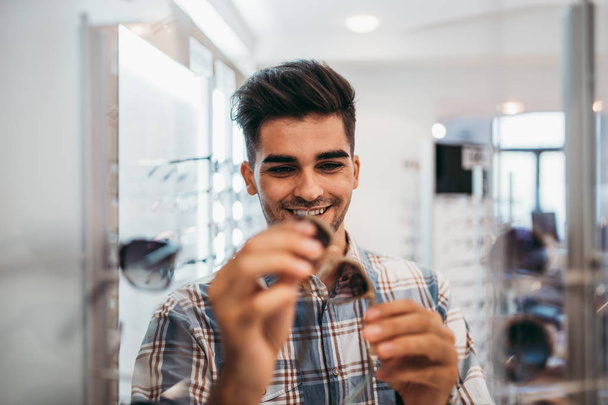 Handsome young man choosing eyeglasses frame in optical store. - Fotó, kép