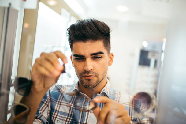 Handsome young man choosing eyeglasses frame in optical store. - Zdjęcie, obraz