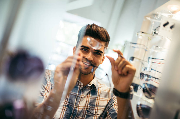 Handsome young man choosing eyeglasses frame in optical store. - Valokuva, kuva