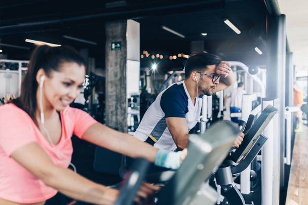 Young and attractive woman and man biking in the fitness gym. They exercising legs and doing cardio workout while riding cycling machines. - 写真・画像