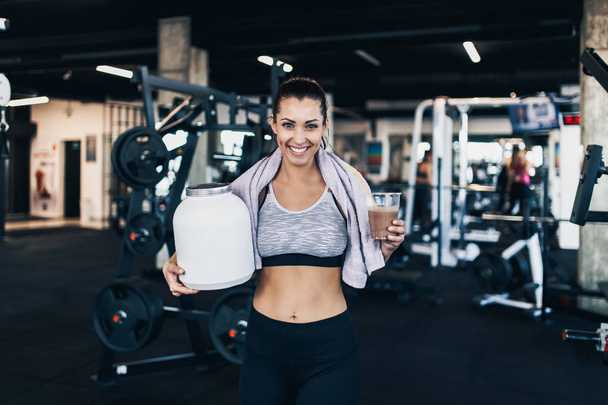 Young attractive woman after successful workout posing and smiling in modern fitness gym while holding big white protein jar and glass of shake with drinking straw in other hand. - Foto, Bild