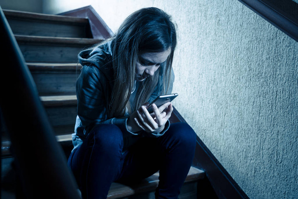 Teenager girl victim of online stalker suffering from cyberbullying abuse feeling lonely and hopeless sitting on stairs with dark light. Dangers of internet, online grooming and harassment concept. - Photo, Image