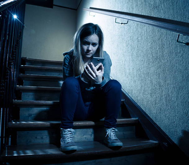 Sad depressed young teenager girl victim of cyberbullying by mobile smart phone siting on stairs feeling lonely, unhappy, hopeless and abused. Bullied by text message on social media app. Dark light - Fotografie, Obrázek