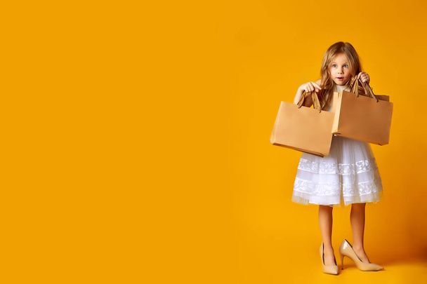 Amazed attractive child in dress and big shoes holding shopping bags on yellow background - Foto, Imagem