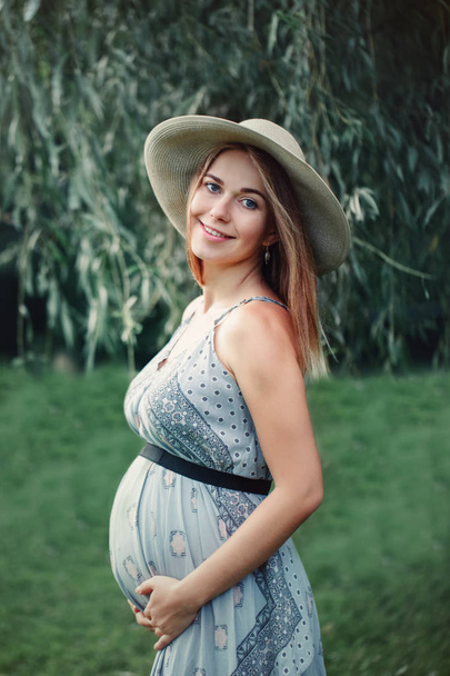 Happy healthy pregnancy and maternity. Portrait of pregnant young Caucasian woman wearing long blue dress and rustic country hat in park outside. Beautiful feminine expecting lady - 写真・画像