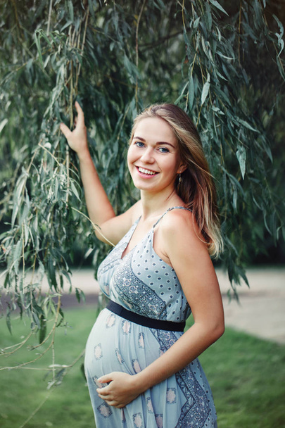 Feliz embarazo saludable y maternidad. Retrato de una joven mujer caucásica embarazada con vestido azul largo posando en el parque exterior. Hermosa sonrisa riéndose femenina esperando dama
 - Foto, imagen