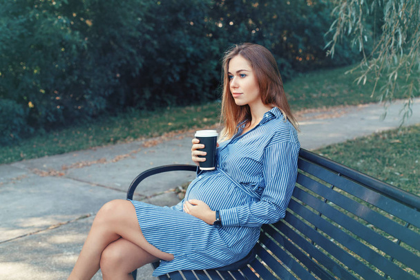 Feliz maternidad sana. Retrato de una joven mujer caucásica embarazada sentada en el banco en el parque afuera y bebiendo café de una taza de papel. Bebida malsana durante el embarazo
 - Foto, Imagen