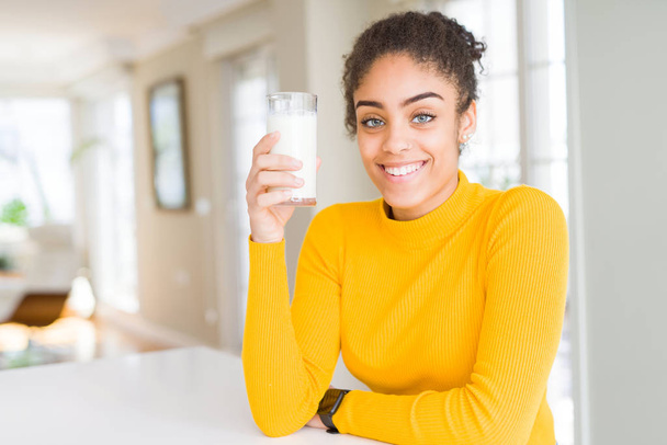 Młoda Afrykańska amerykańska kobieta pijąc szklankę świeżego mleka z szczęśliwą twarzą i uśmiechając się z pewnym uśmiechem, pokazując zęby - Zdjęcie, obraz