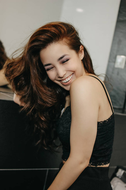 Felice sensuale sorridente asiatica giovane donna con lunghi capelli ricci scuri davanti allo specchio. Ragazza divertirsi alla festa
 - Foto, immagini