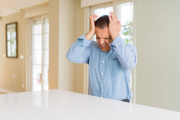 Hombre de mediana edad sentado en casa sufriendo de dolor de cabeza desesperado y estresado porque el dolor y la migraña. Manos en la cabeza
. - Foto, Imagen