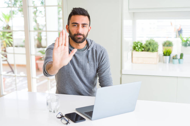 Handsome hispanic man working using computer laptop with open hand doing stop sign with serious and confident expression, defense gesture - Fotó, kép