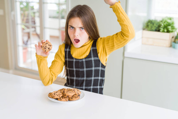 Krásné mladé děvče, které žere čokoládové hranolky sušenky otrávené a frustrované křikem hněvem, šílením a křikem s zvedanou rukou, koncept hněvu - Fotografie, Obrázek