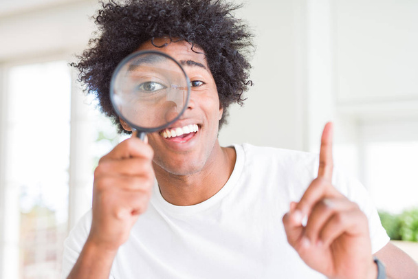 Αφρικανική αμερικανική άνθρωπος που ψάχνει μέσα από μεγεθυντικό γυαλί εξέπληξε με μια ιδέα ή θέμα τεντωμένο με χαρούμενο πρόσωπο, αριθμός - Φωτογραφία, εικόνα