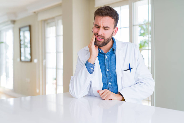 Handsome doctor man wearing medical coat at the clinic touching mouth with hand with painful expression because of toothache or dental illness on teeth. Dentist concept. - 写真・画像
