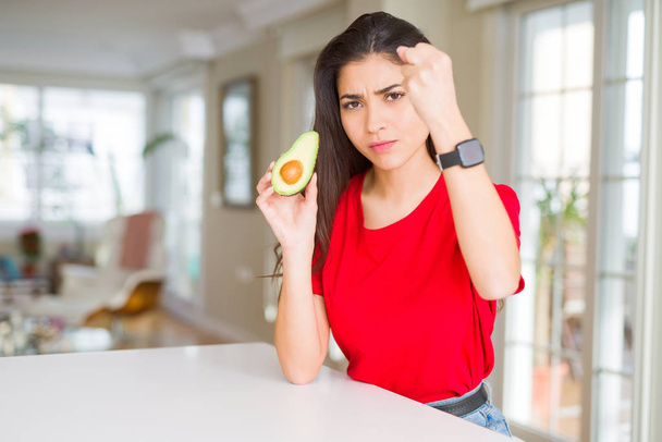 Young woman eating healthy avocado annoyed and frustrated shouting with anger, crazy and yelling with raised hand, anger concept - Foto, afbeelding