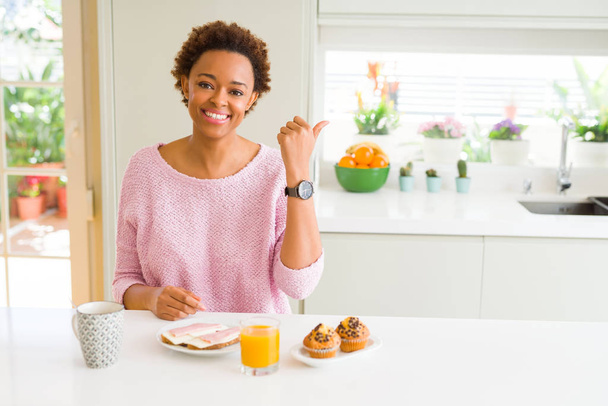Молода жінка їсть breaksfast в першій половині дня вдома посміхається з щасливим обличчям, дивлячись і вказуючи на сторону з великим пальцем. - Фото, зображення
