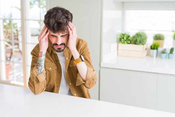 Hombre joven con chaqueta casual sentado en la mesa blanca que sufre de dolor de cabeza desesperado y estresado porque el dolor y la migraña. Manos en la cabeza
. - Foto, imagen