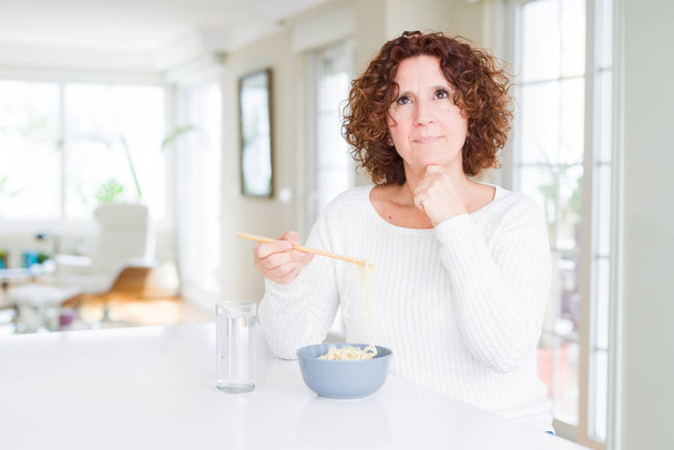 Senior woman eating asian noodles using chopsticks serious face thinking about question, very confused idea - Foto, Imagem