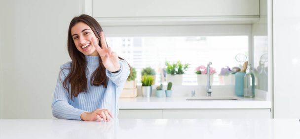 Широкоугольная фотография красивой молодой женщины, сидящей дома на белом столе, улыбающейся, глядя в камеру и показывающей пальцы, делающие знак победы. Номер два:
. - Фото, изображение