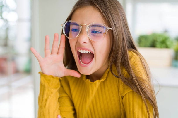 Beautiful young girl kid wearing glasses shouting and screaming loud to side with hand on mouth. Communication concept. - Фото, изображение