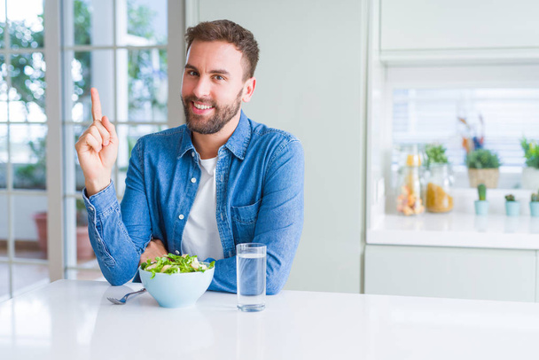 Красивый мужчина ест свежий здоровый салат с большой улыбкой на лице, указывая рукой и пальцем в сторону, глядя в камеру
. - Фото, изображение