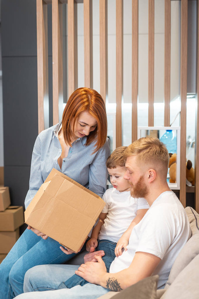 Young happy family with kid unpacking boxes together sitting on sofa - Photo, Image