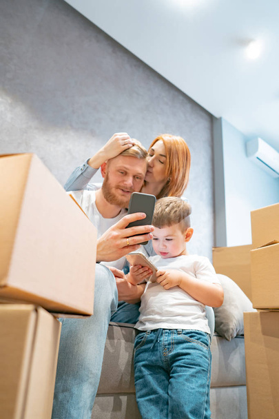 Young happy family with kid unpacking boxes together sitting on sofa - Photo, Image
