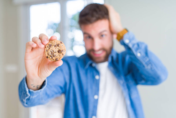 Knappe man eten chocolade chips cookies met benadrukt de hand op het hoofd, geschokt met schaamte en verrassing gezicht, boos en gefrustreerd. Angst en boos voor fout. - Foto, afbeelding