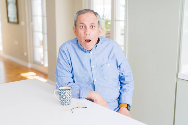 Handsome senior man drinking a cup of coffee at home afraid and shocked with surprise expression, fear and excited face. - Photo, Image