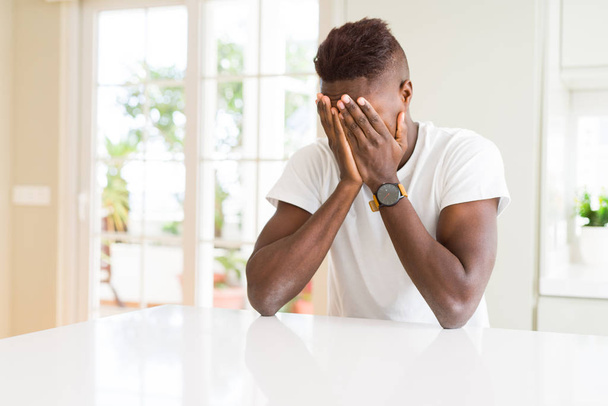 Bel homme afro-américain sur une table blanche à la maison avec une expression triste couvrant le visage des mains tout en pleurant. Concept de dépression
. - Photo, image