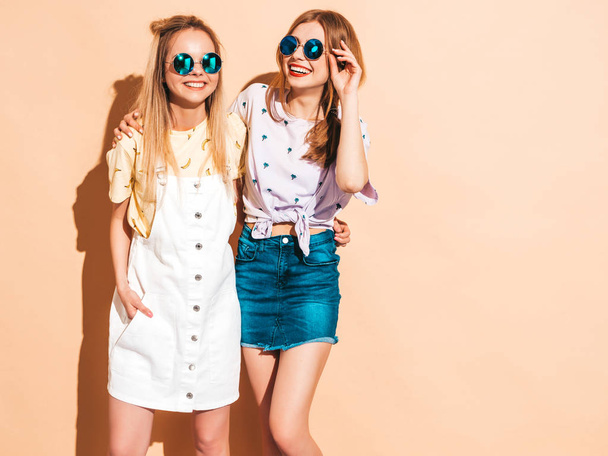 Dwie młode piękne, uśmiechnięte dziewczyny blond hipster w modne letnie kolorowe koszulki odzież. Sexy kobiety beztroski, pozować w pobliżu beżowej ściany w okulary przeciwsłoneczne. Pozytywnych wzorców zabawy - Zdjęcie, obraz
