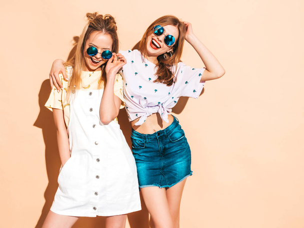Two young beautiful smiling blond hipster girls in trendy summer colorful T-shirt clothes. Sexy carefree women posing near beige wall in round sunglasses. Positive models having fun - Foto, Imagem
