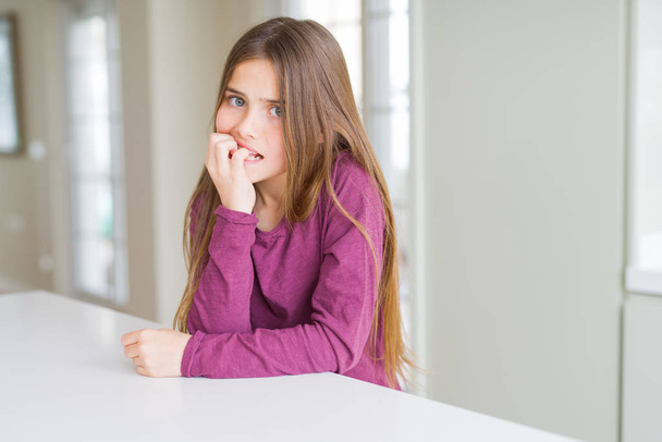 Mooie jonge meisje Kid op witte tafel op zoek gestrest en nerveus met de handen op de mond bijten nagels. Angst probleem. - Foto, afbeelding