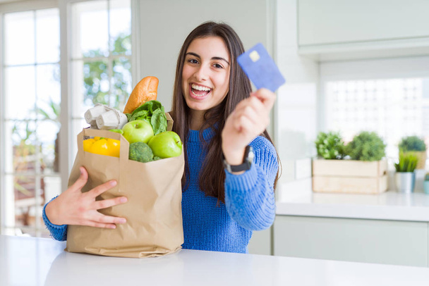 Young woman holding a paper bag full of fresh groceries and showing credit card as payment metod - Fotó, kép