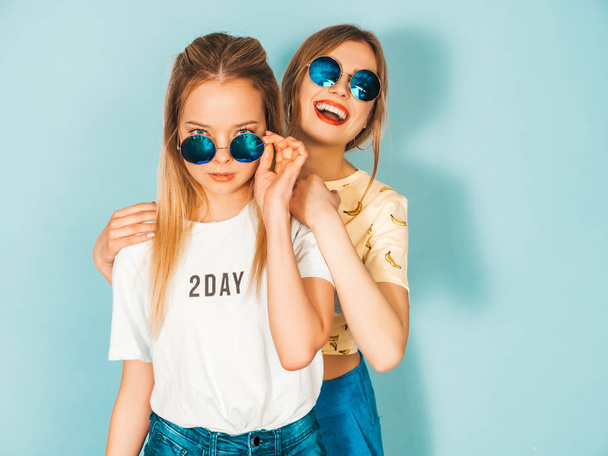 Two young beautiful smiling blond hipster girls in trendy summer colorful T-shirt clothes. Sexy carefree women posing near blue wall in round sunglasses. Positive models having fun - Foto, Imagem