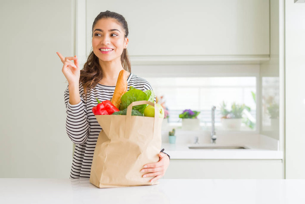 Jeune femme tenant sac en papier plein d'épicerie très heureux pointant avec la main et le doigt sur le côté
 - Photo, image