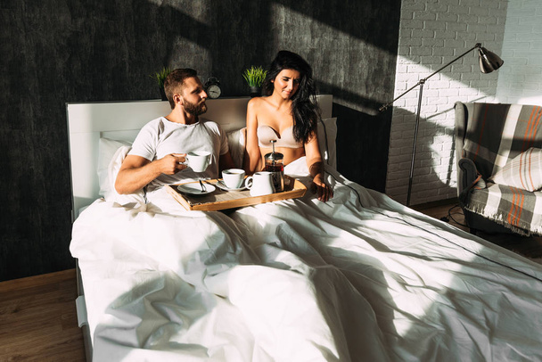 Beautiful couple in bed. Lovers in bed. Happy loving couple having breakfast in bed. Family relationships. Young couple drinking tea. Man and woman having Breakfast in their bedroom - Fotoğraf, Görsel